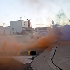 Color photograph of plumes of orange and purple smoke rising alongside a black and yellow barrier on a paved street and sidewalk