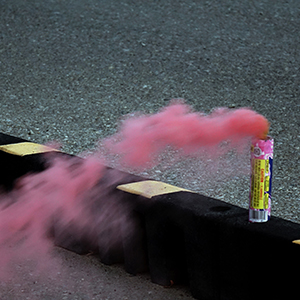 Color photograph of a plume of pink smoke emerging from a pink smoke canister perched on a black and yellow barrier