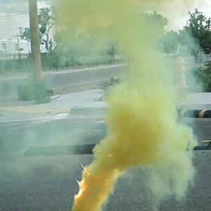 Color photograph of a plume of yellow smoke and sparks rising from a yellow smoke canister perched on a black and yellow barrier