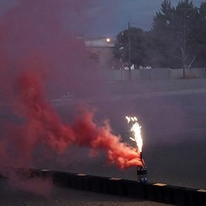 Color photograph of red smoke and sparks rising from smoke canisters perched on a black and yellow barrier at dusk