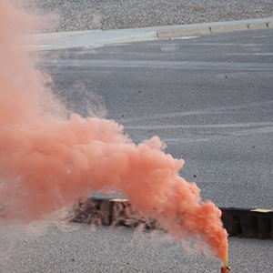 Color photograph of orange smoke rising from a canister perched on a black and yellow barrier