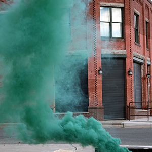 Color photograph of green smoke rising from a black and yellow barrier on a paved street in front of a brick building