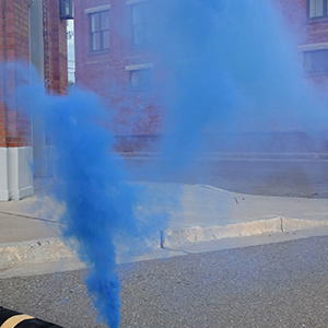 Color photograph of blue smoke rising from a canister alongside a black and yellow barrier on a paved street in front of a brick building