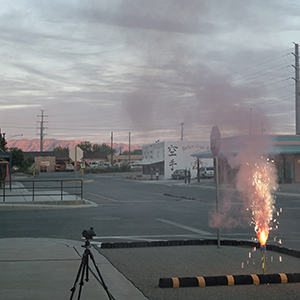 Color photograph of sparks and smoke rising from a torch-like support inside a black and yellow barrier on a paved street with a camera on a tripod nearby and buildings in the distance