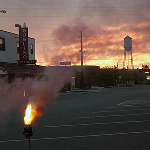 Color photograph of sparks and smoke rising from a torch-like support on a paved street with a water tower in the distance at dusk