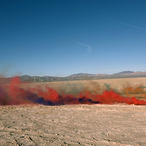 Color photograph of plumes of red and purple smoke emanating from the desert floor