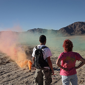 Color photograph of Rusty Johnson and Judy Chicago standing in the desert with their backs to the camera watching plumes of orange and green smoke