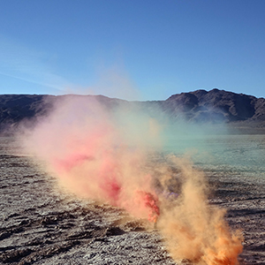 Color photograph of plumes of orange, pink, and green smoke rising from the desert floor