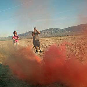 Color photograph of Judy Chicago and Chris Souza, taking a picture, and standing in the desert looking at plumes of red and yellow smoke