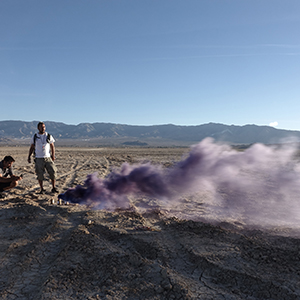 Color photograph of Chris Souza, kneeling and taking a picture, and Rusty Johnson, standing, in the desert looking at a plume of purple smoke