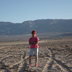 Color photograph of Judy Chicago standing in the desert and watching a plume of white smoke